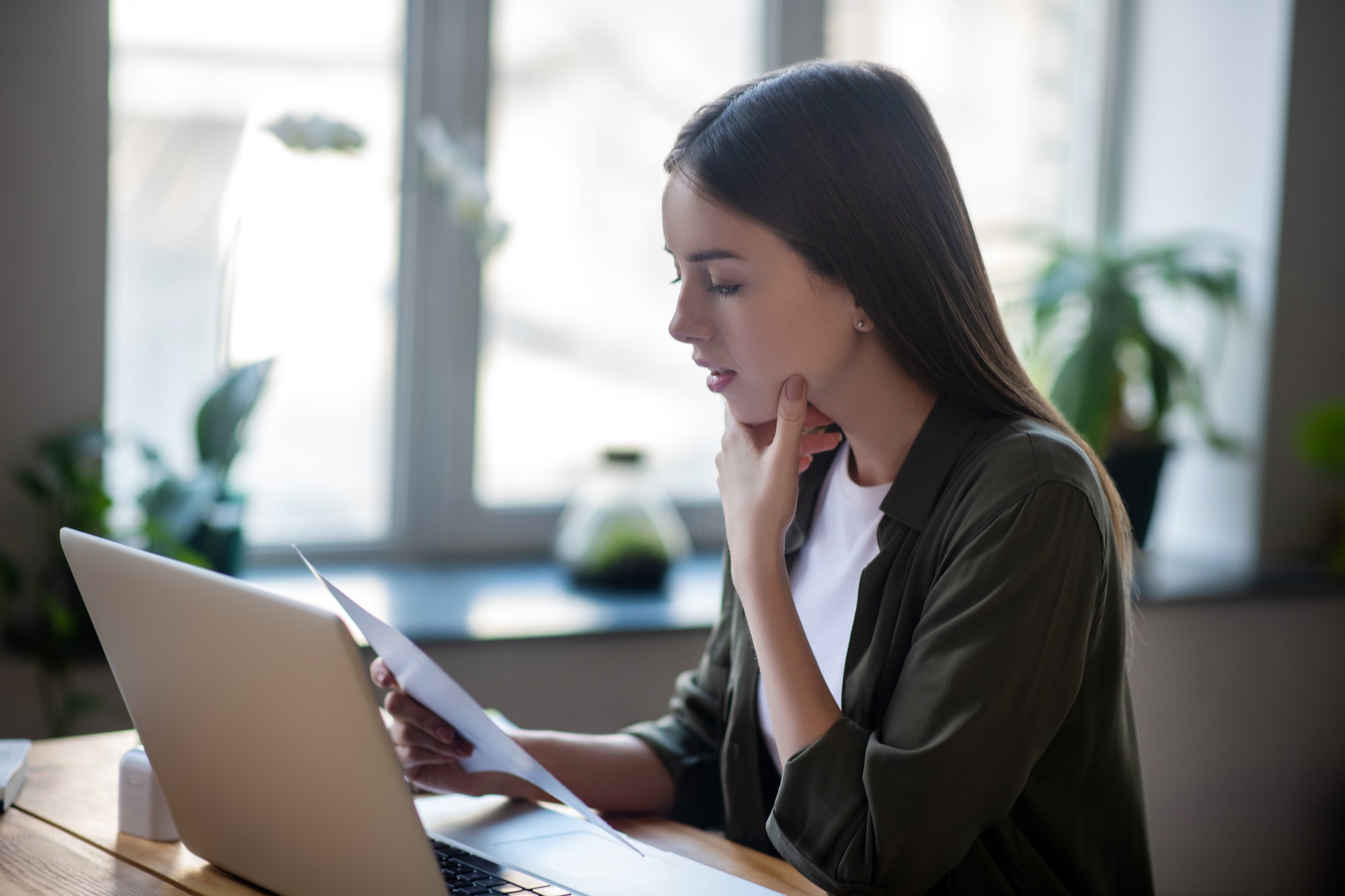 Girl sitting in front of laptop carefully looking at paper document